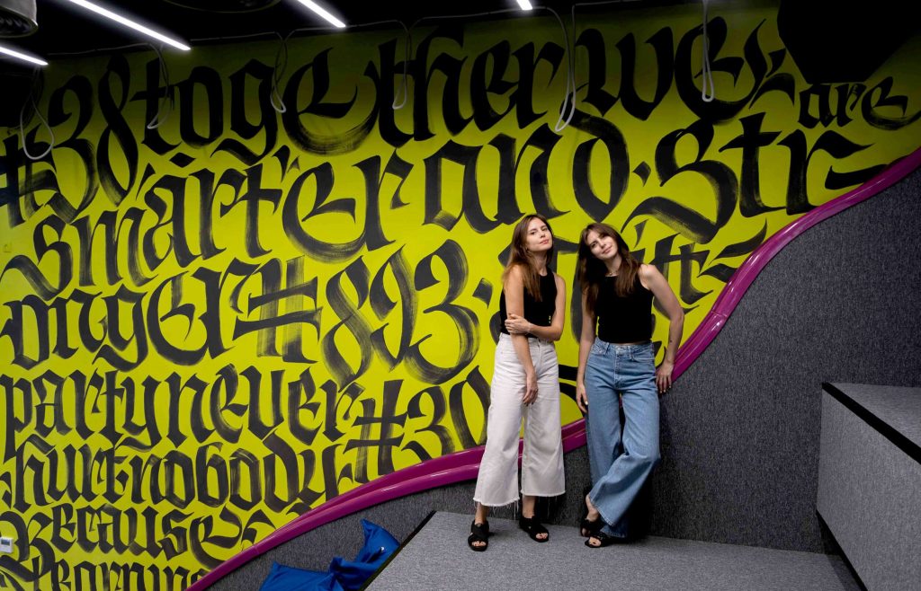 Die Typografinnen von Vikavita vor ihrem Kunstwerk der Schriftmalerei – schwarze Schrift auf gelbem Grund